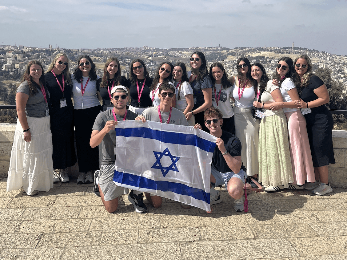 Group of young adults posing on a overlook holding an Israeli flag, with a cityscape in the background.