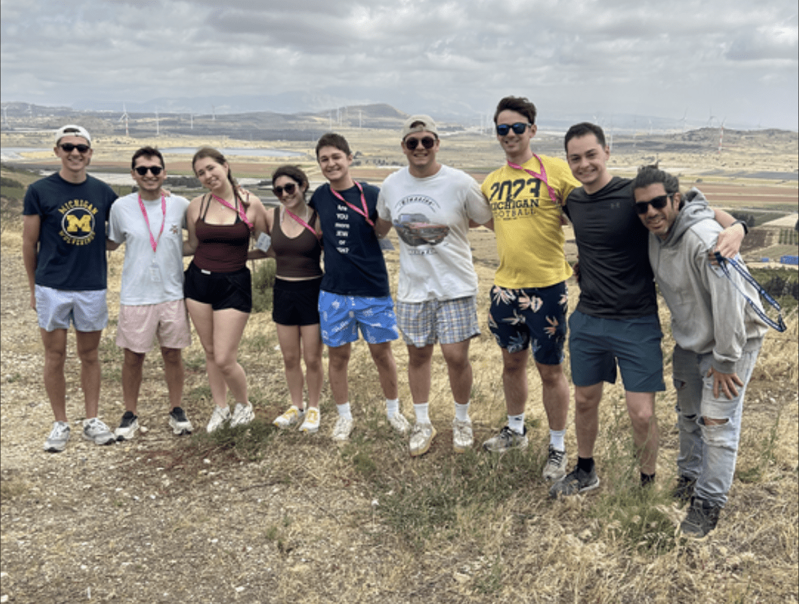Group of ten friends posing together on a dry hillside with a wide rural landscape behind them, smiling and wearing casual summer clothing.